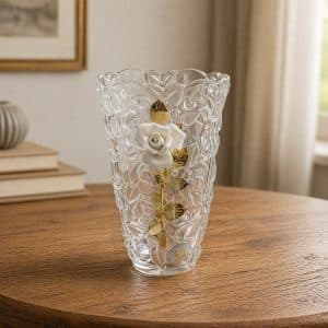 Crystal vase with a white rose and gold leaves, sitting on a wooden table in a softly lit room.