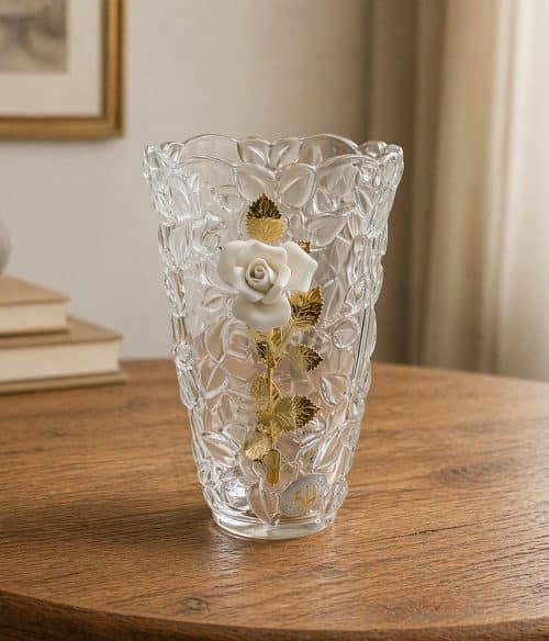 Crystal vase with a white rose and gold leaves, sitting on a wooden table in a softly lit room.