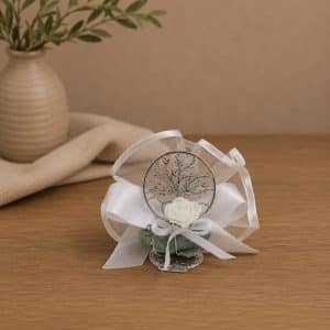 Wedding boutonniere with a white rose, silver tree charm, and satin ribbons on a wooden table.