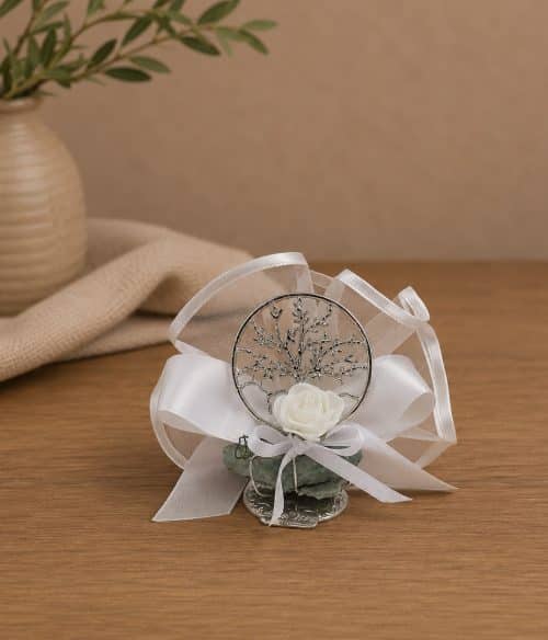 Wedding boutonniere with a white rose, silver tree charm, and satin ribbons on a wooden table.
