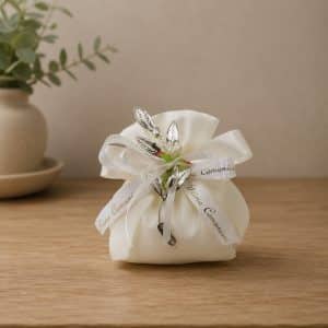 White satin favor bag tied with ribbons and silver leaf charms, a First Communion keepsake on a wooden surface