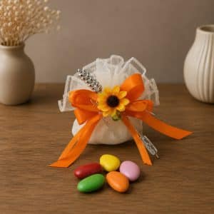 Decorative favor: white organza bag tied with orange ribbon, accented by a yellow sunflower and silver pins, placed on a wooden table with colorful candies nearby.