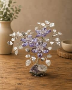 Decorative crystal tree sculpture with purple gem flowers and silver leaves on a wooden table, beige vase blurred in background.