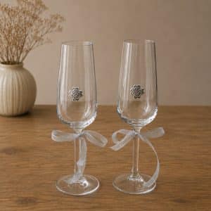 Two clear champagne flutes with silver ribbon bows on the stems, each featuring a small metal flower emblem, set on a wooden table.