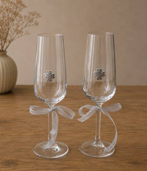 Two clear champagne flutes with silver ribbon bows on the stems, each featuring a small metal flower emblem, set on a wooden table.
