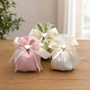 Three satin gift pouches in pink, green, and cream with ivory ribbons and heart-shaped clasps arranged on a wooden table, likely decorative favor bags.