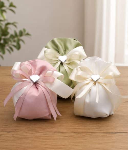 Three satin gift pouches in pink, green, and cream with ivory ribbons and heart-shaped clasps arranged on a wooden table, likely decorative favor bags.