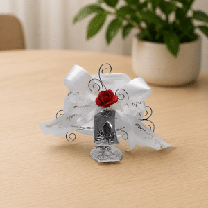 Silver metal wedding favor with white ribbons and a red rose accent on a light wooden table.