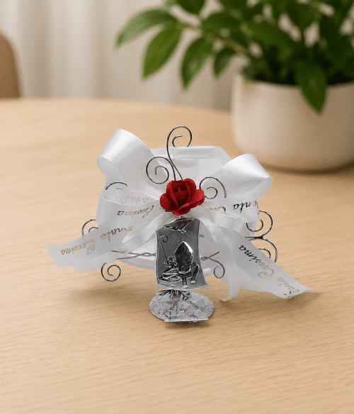 Silver metal wedding favor with white ribbons and a red rose accent on a light wooden table.