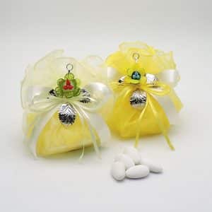 Two yellow organza gift pouches tied with white ribbons, each with colorful flower charms and metallic leaf accents, on a white surface with white almonds nearby.