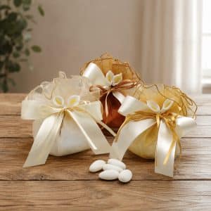 Gift pouches with ivory and gold ribbons on a wooden table, decorative sachets with white flowers and sugar-coated almonds nearby.