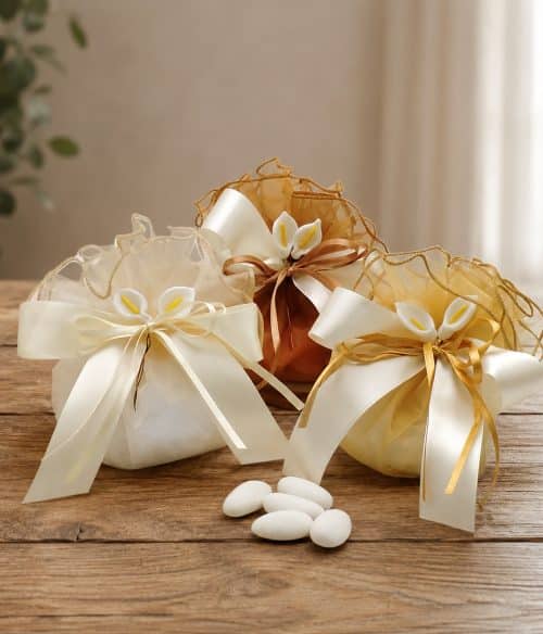Gift pouches with ivory and gold ribbons on a wooden table, decorative sachets with white flowers and sugar-coated almonds nearby.